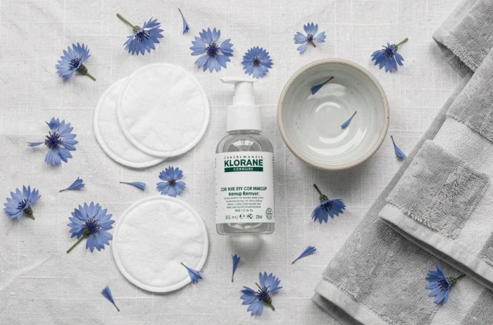 Flatlay du démaquillant yeux Klorane au bleuet avec cotons et pétales de bleuet sur fond blanc